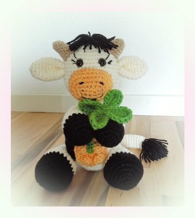White and black crocheted cow amigurumi sitting and holding a green crochet clover and a small orange decorative detail