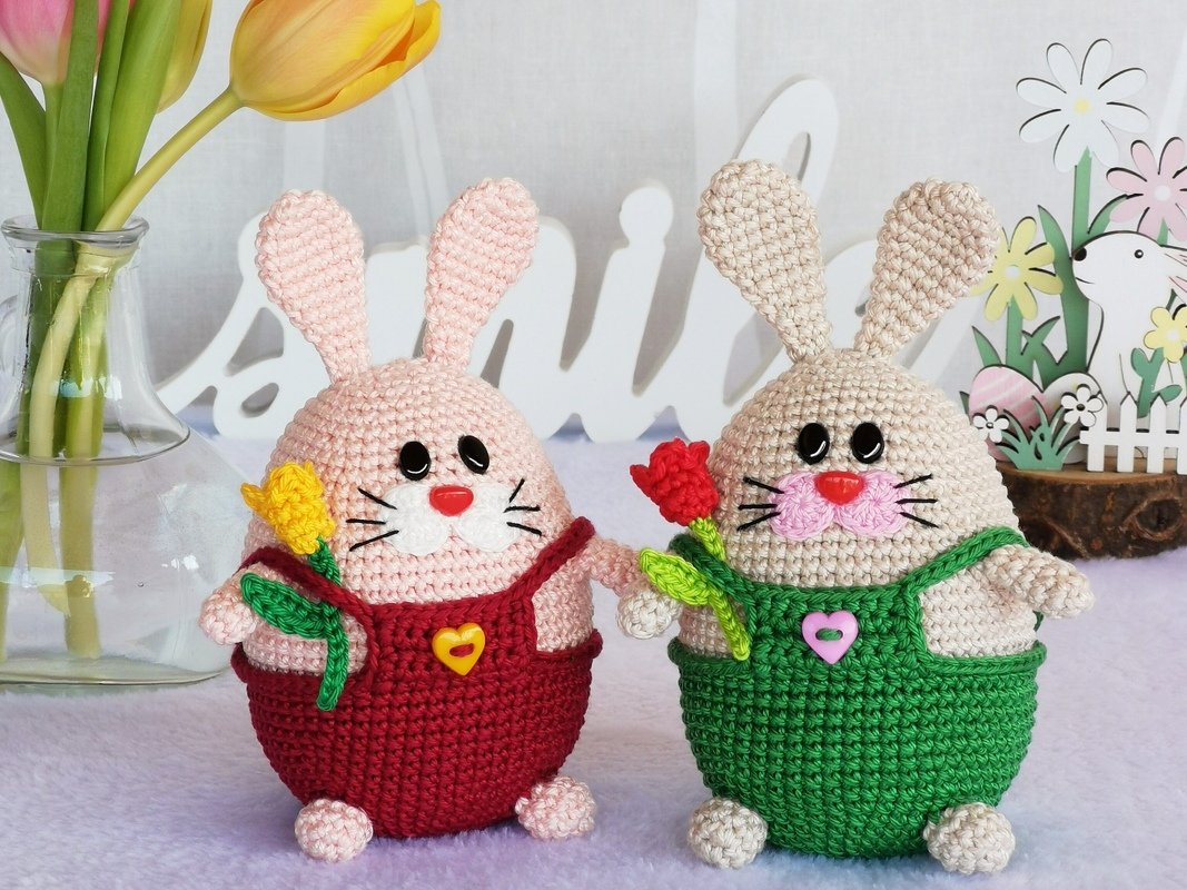 Pair of crocheted amigurumi bunnies: pink in red overalls with a yellow flower, and cream in green overalls with a red flower, on a spring backdrop.