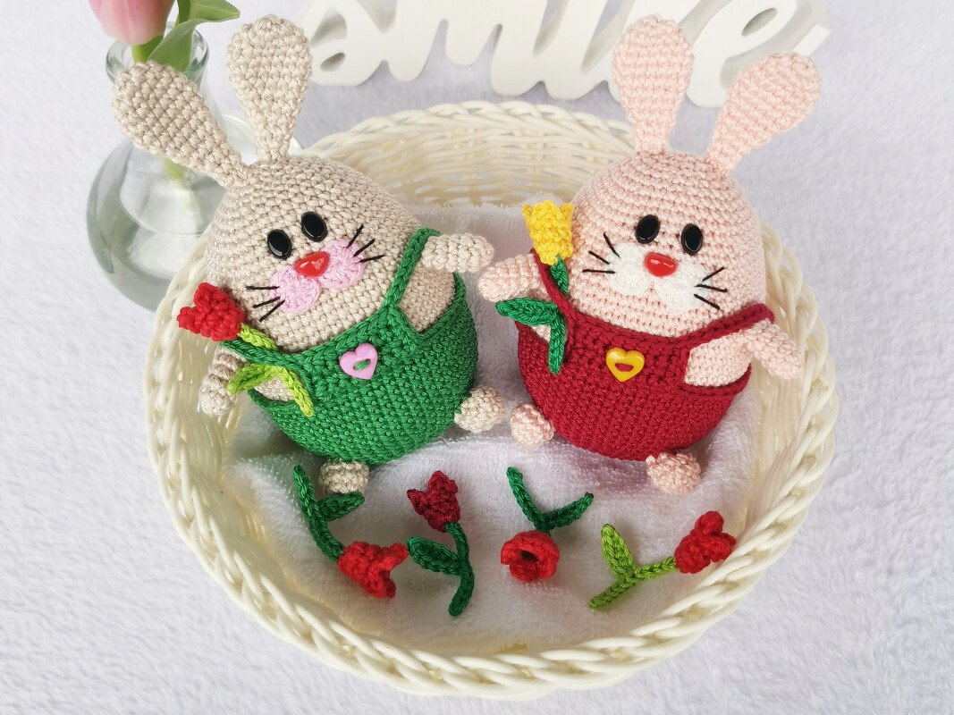 Two crocheted amigurumi rabbits in a white basket, wearing green and red outfits and holding small flowers