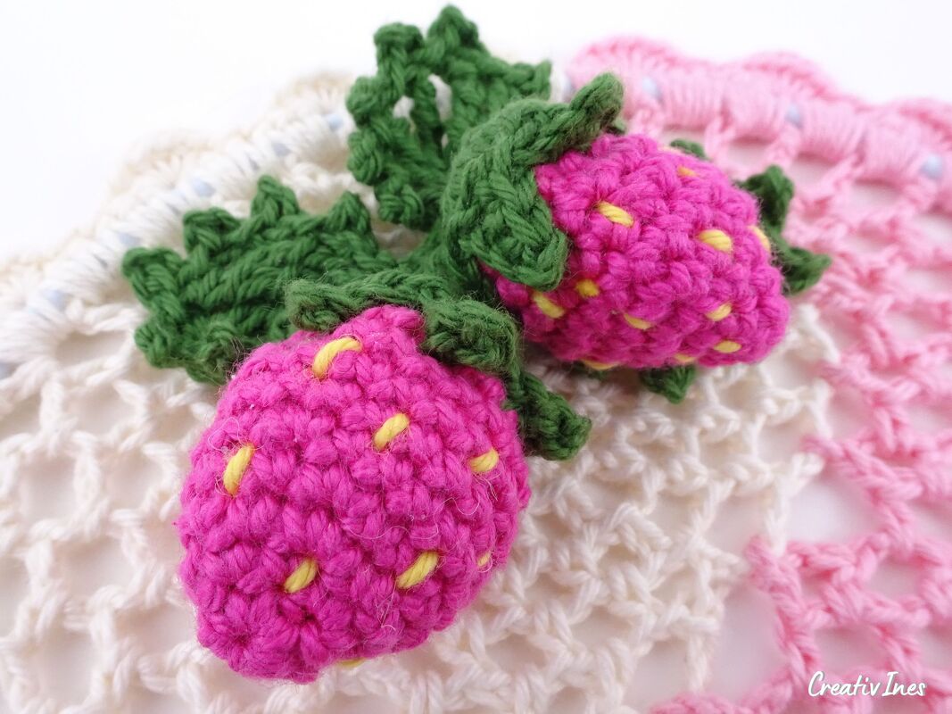 Zwei pinke gehäkelte Erdbeeren mit gelben Stichen und grünen Blättern auf heller gehäkelter Unterlage
