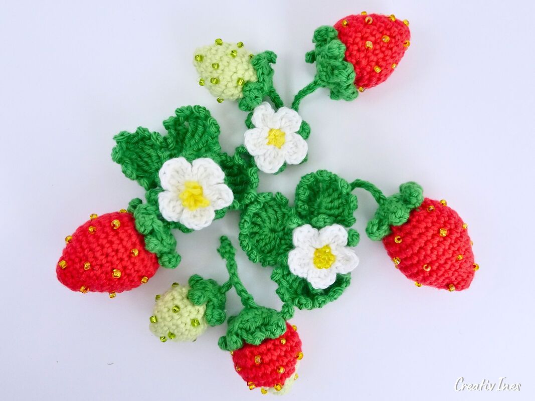 Gehäkelte rote und hellgrüne Erdbeeren, grüne Blätter und weiße Blüten mit Perlen auf hellem Hintergrund