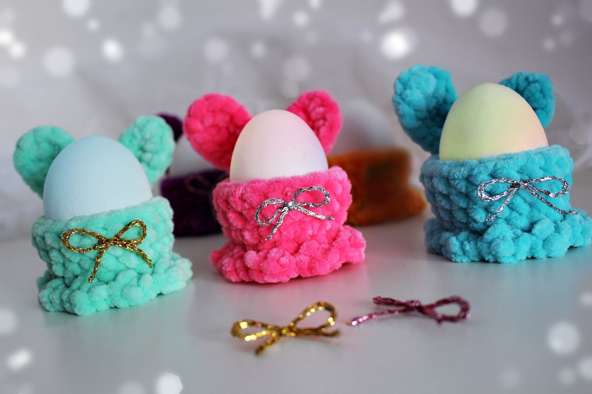 Nahaufnahme von drei gehäkelten, flauschigen Eierwärmern in Mint, Pink und Türkis mit Öhrchen und Schleifen, die Eier halten.