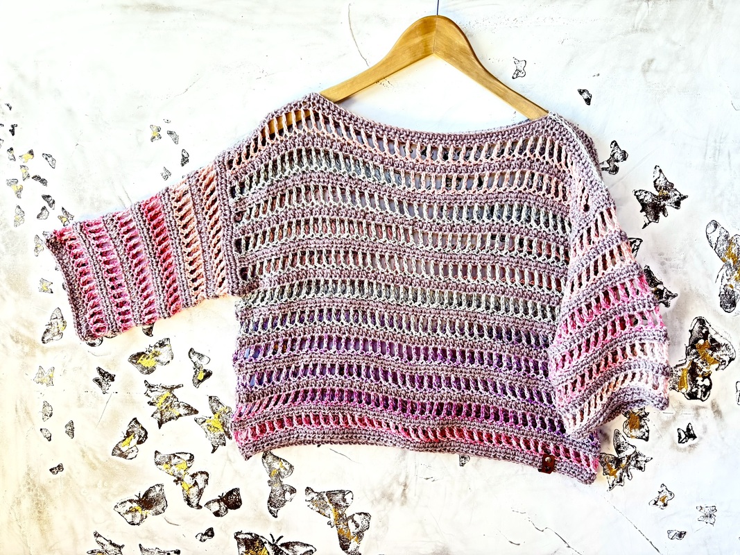 Gehäkeltes Damen-Shirt mit Lochmuster und pastellfarbenem Farbverlauf auf einem Holzbügel