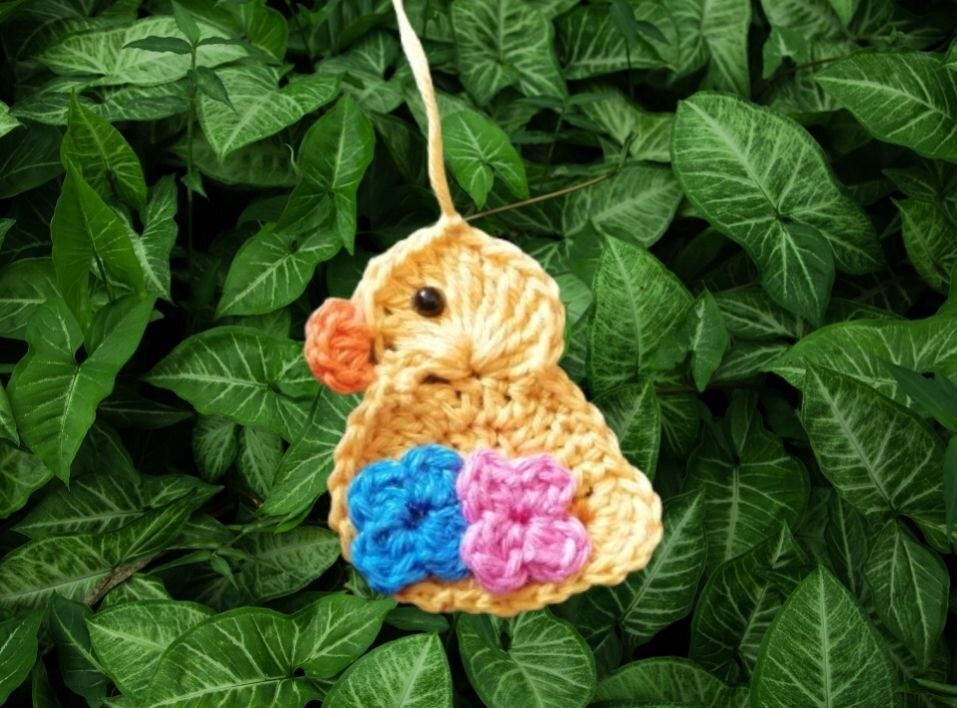Gehäkeltes gelbes Küken-Anhänger mit blauen und rosa Blüten vor grünen Blättern