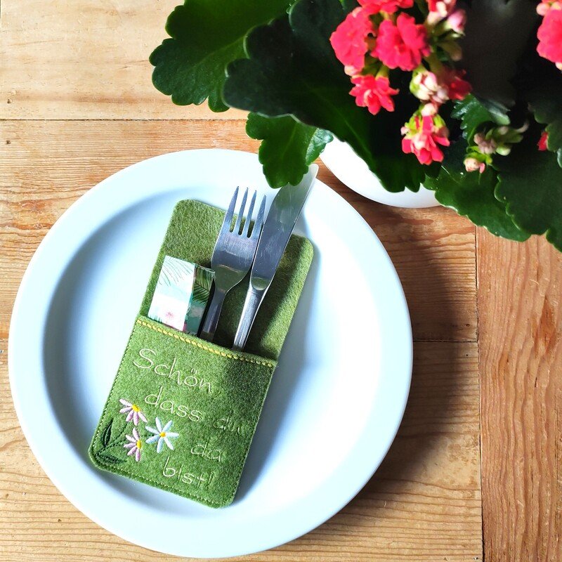 Grüne Filz-Bestecktasche mit gesticktem Schriftzug "Schön, dass du da bist!" und Blumen, mit Besteck auf weißem Teller