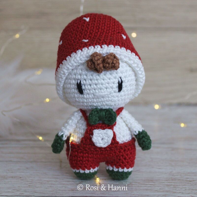 Gehäkeltes Glückspilz‑Amigurumi mit roter Mütze, grüner Fliege und roten Latzhosen vor hellem Hintergrund