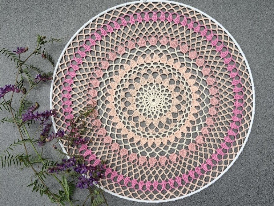 Rundes gehäkeltes Mandala in Rosa- und Beigetönen in weißem Reifen, links violette Blütenzweige