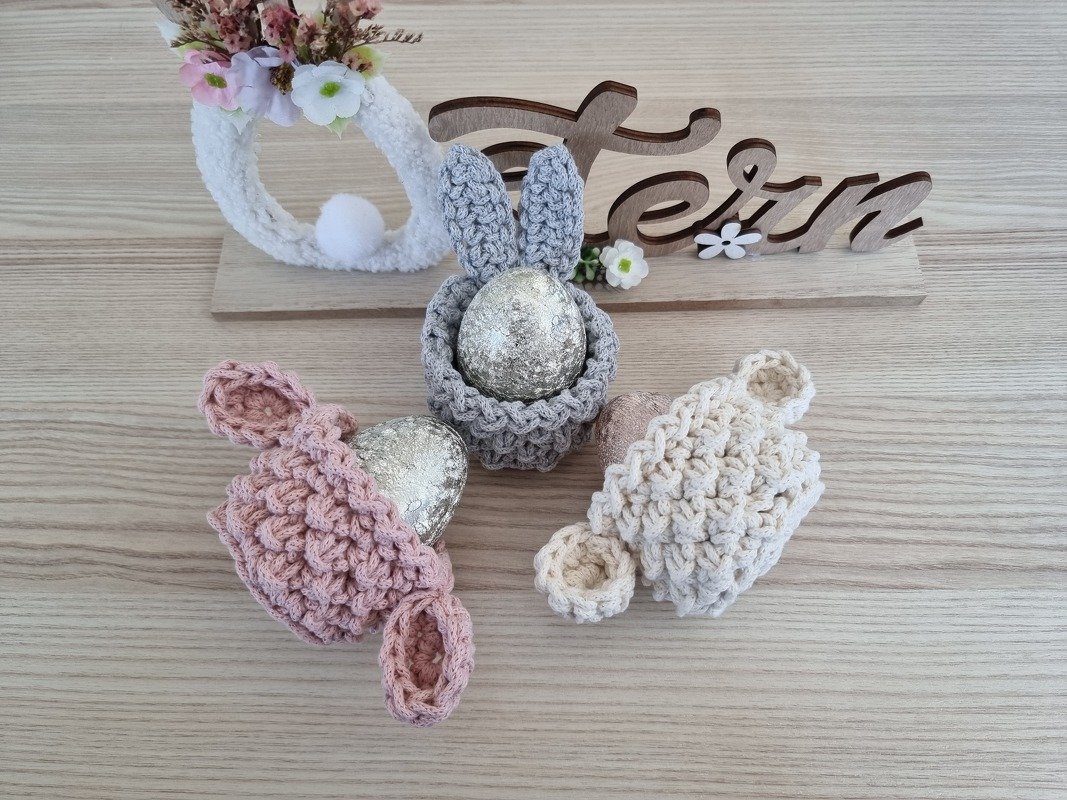 Drei gehäkelte Hasen-Eierwärmer in Grau, Rosa und Creme mit dekorativen Ostereiern auf einem Holztisch