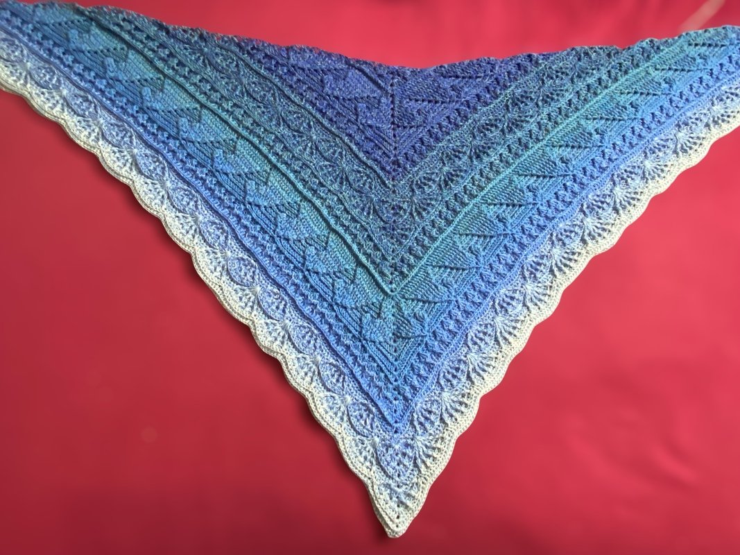 Blue-to-white gradient crochet triangle shawl with lace motifs and scalloped edge on red background
