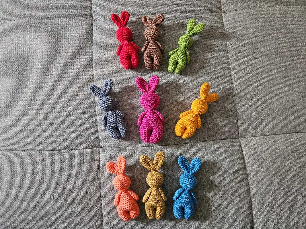 Neun gehäkelte Amigurumi-Hasen in bunten Garnfarben auf grauem Stoff