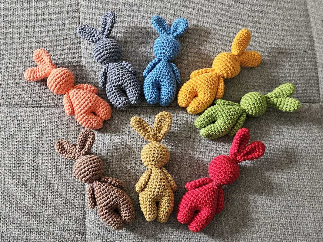 Seven small crocheted amigurumi bunnies in assorted colors arranged in a circle on grey fabric.