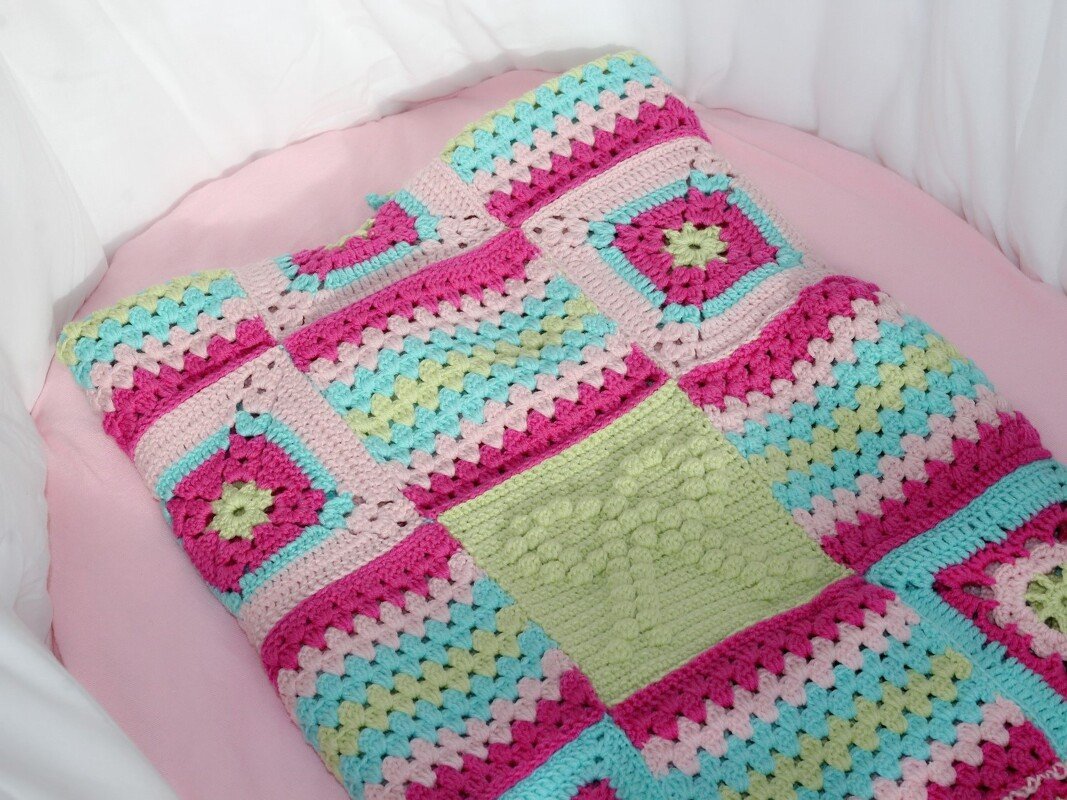 Bunte gehäkelte Babydecke aus verschiedenen Quadraten (Granny Squares und Noppenmuster) in Rosa, Pink, Türkis und Hellgrün auf einem rosa Laken.