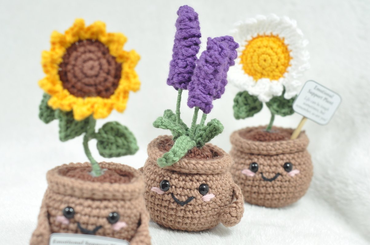 Close-up of three smiling crocheted pots holding a sunflower, lavender and daisy, showing stitch texture and bead eyes