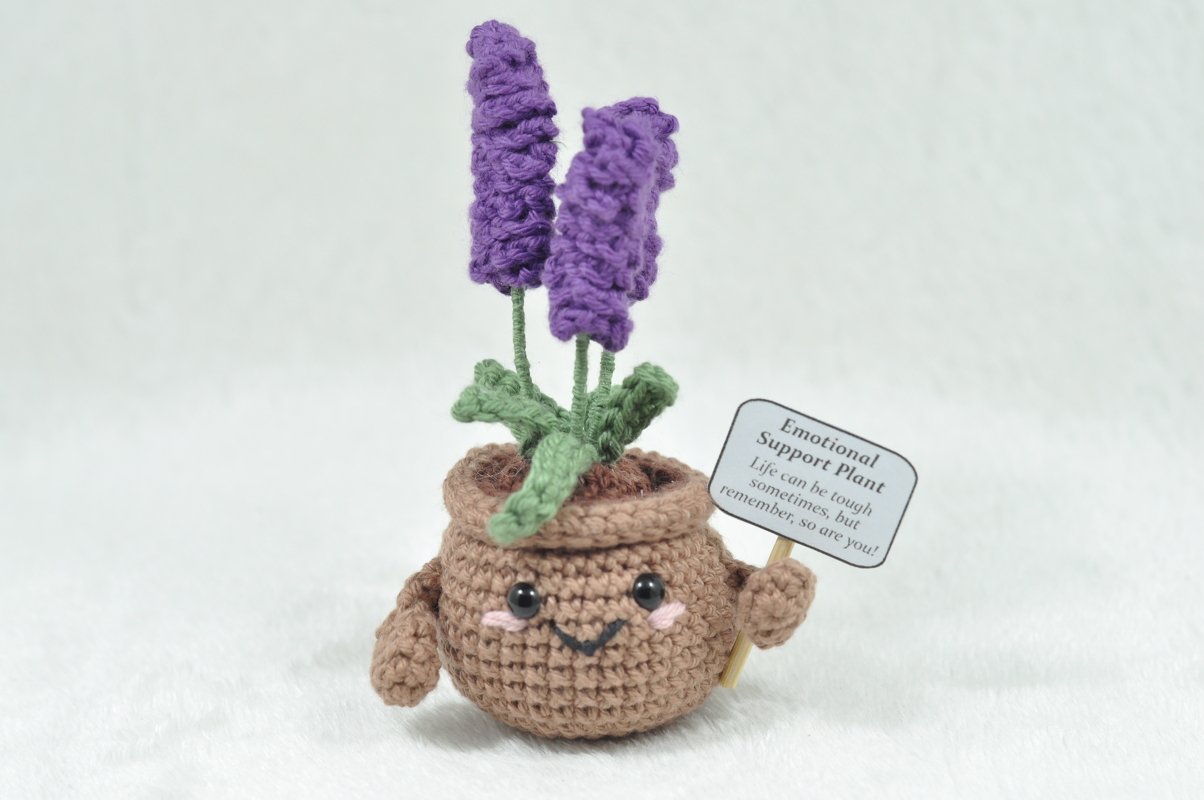 Gehäkelter brauner Blumentopf mit zwei violetten Lavendelblüten, grünen Blättern, lachendem Gesicht und kleinem Schild