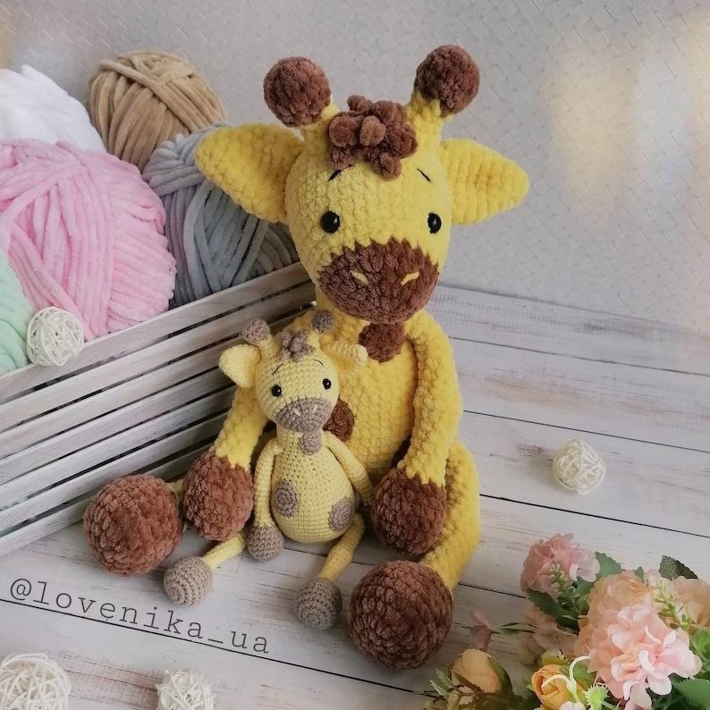 Two handmade yellow and brown crocheted amigurumi giraffes beside a crate of colorful yarn skeins on a white wooden surface.