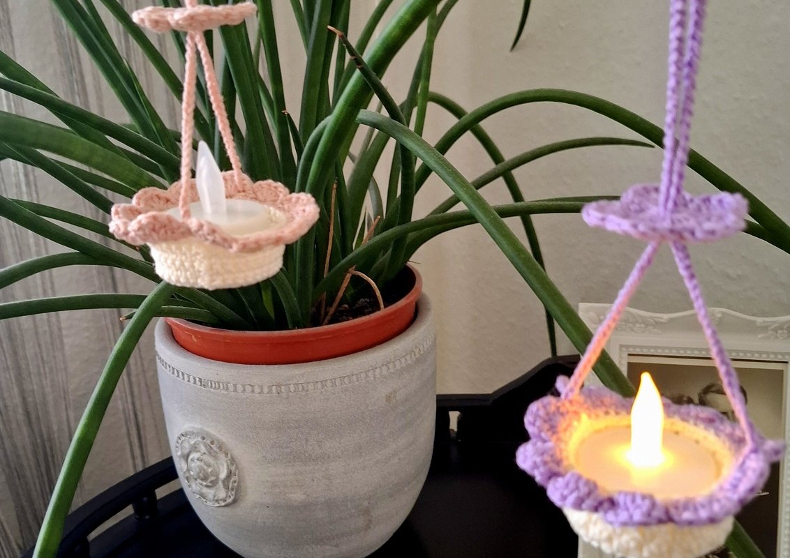 Nahaufnahme zweier schnell gehäkelter Hänge-Teelichthalter mit LED-Teelichtern, eine rosa und eine lila, hängend vor einer Zimmerpflanze