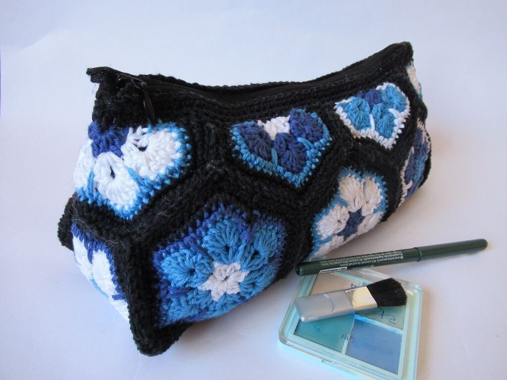 Gehäkelte Kosmetiktasche aus blauen, weißen und schwarzen African‑Flower‑Hexagonen mit Reißverschluss, daneben Eyeliner und Lidschatten