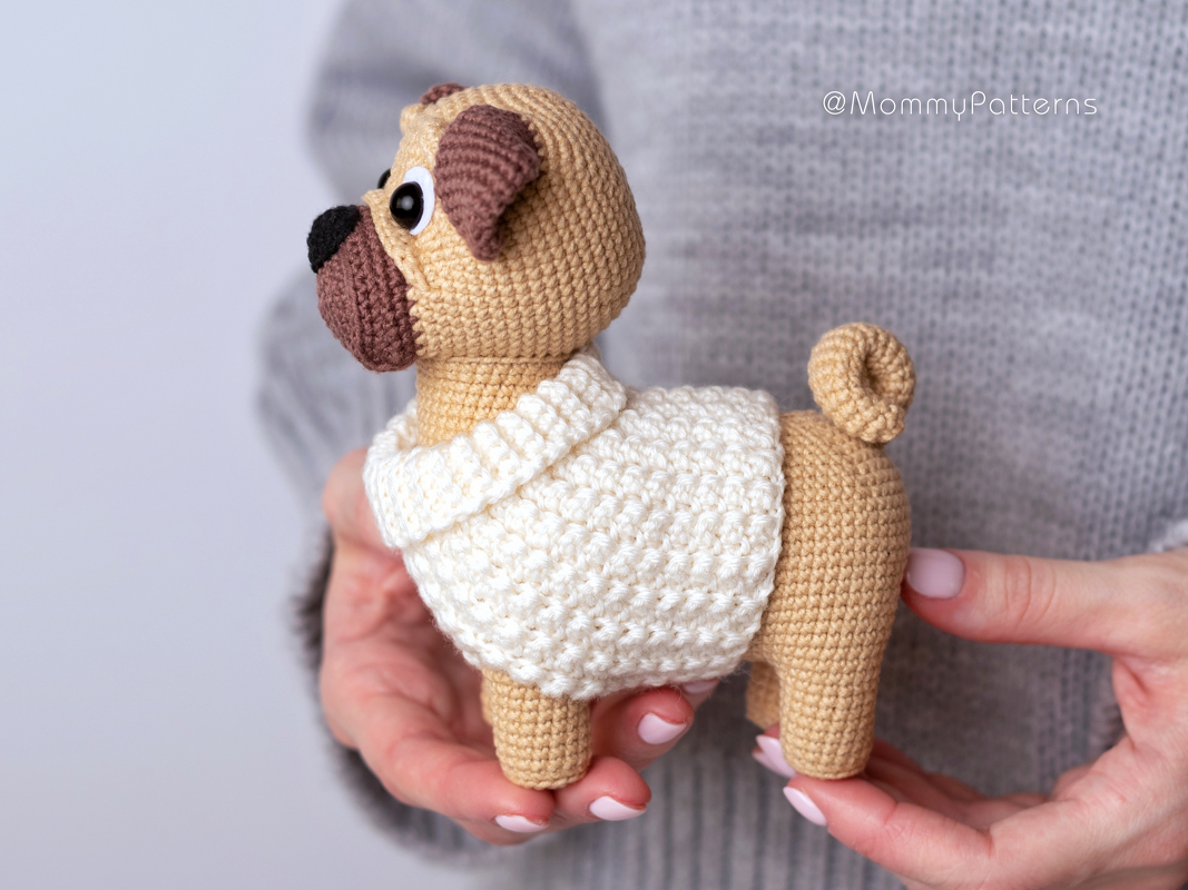Beige crocheted pug amigurumi wearing a cream sweater held in hands.