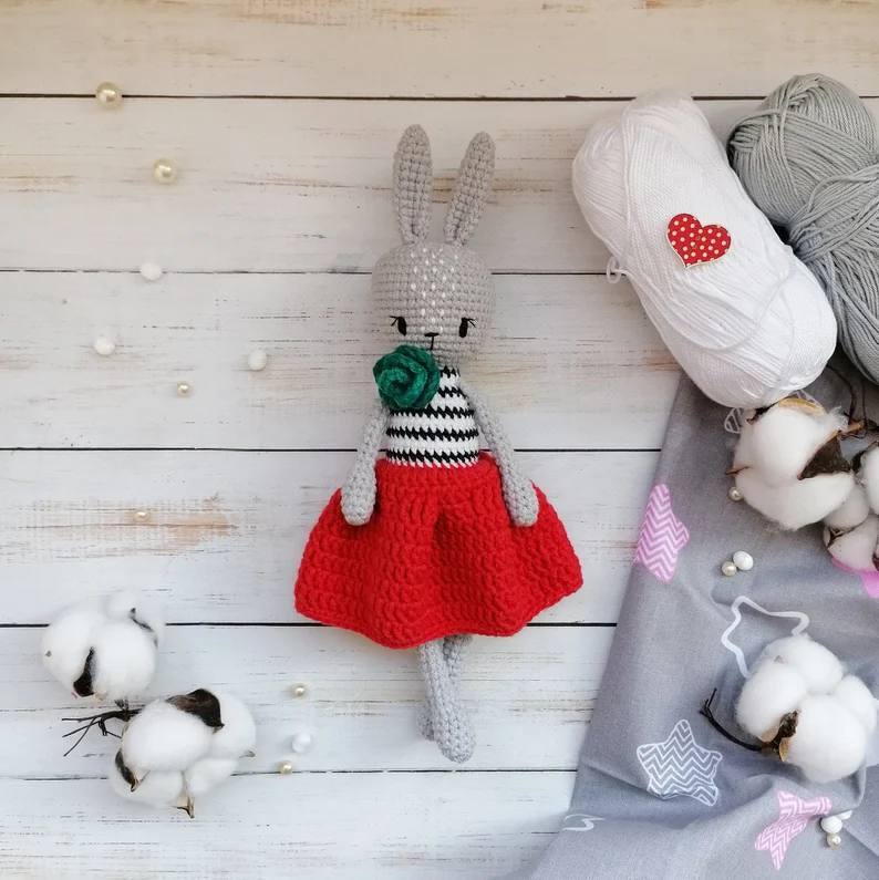 Grey crocheted amigurumi rabbit wearing a red skirt and striped top, shown with yarn and cotton.