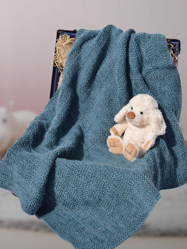 Gestrickte blaue Babydecke mit dezentem Strukturmuster neben einem hellen Plüschtier in einer Holzkiste
