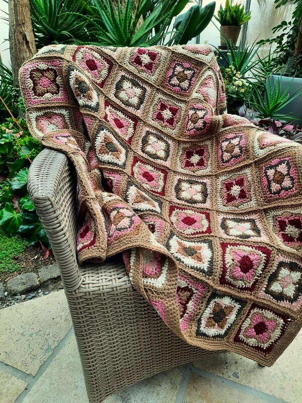 Boho-Häkeldecke aus Granny-Squares in Braun- und Rosatönen über einem Korbsessel drapiert