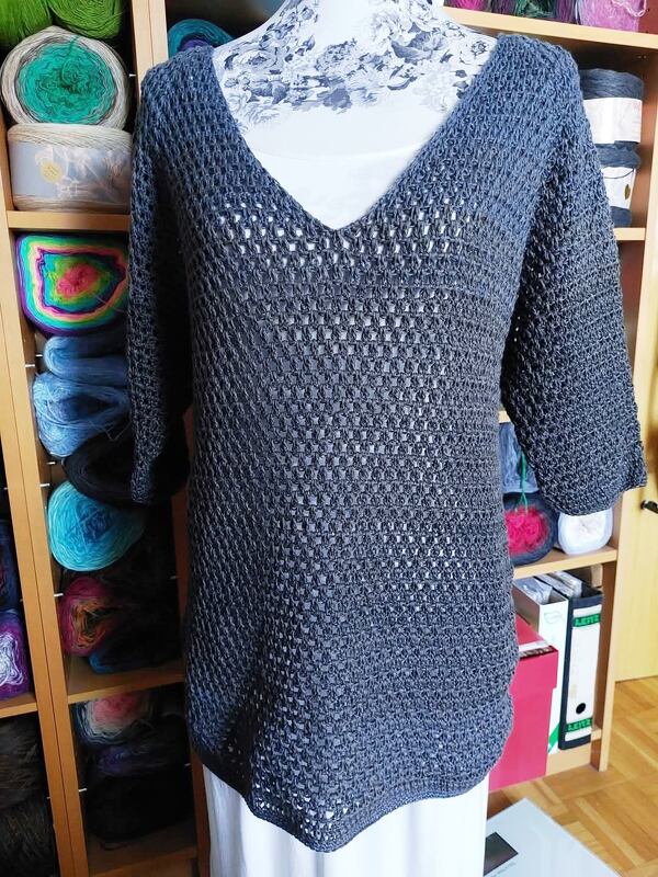 Mannequin trägt graue, locker gehäkelte Tunika mit V-Ausschnitt und 3/4-Ärmeln vor Regal mit Garnrollen