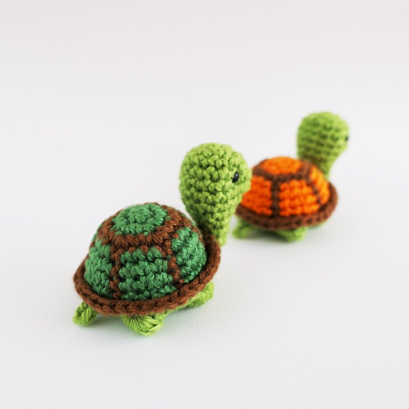 Zwei handgehäkelte Amigurumi‑Schildkröten mit grün‑braunem und orange‑braunem Panzer auf hellem Hintergrund.