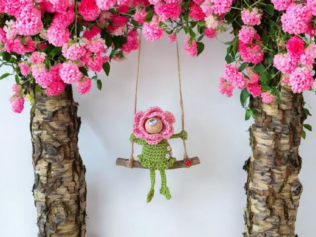 Gehäkelte Blumenfigur mit rosa Blütenkopf und grünem Körper auf Holzschaukel unter rosa Blüten.