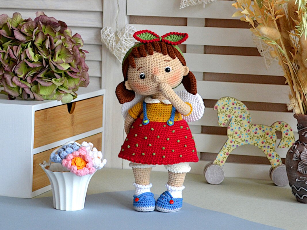Handmade crocheted amigurumi girl doll wearing a red bobble skirt, yellow top, blue shoes and a leaf headband, standing on a table