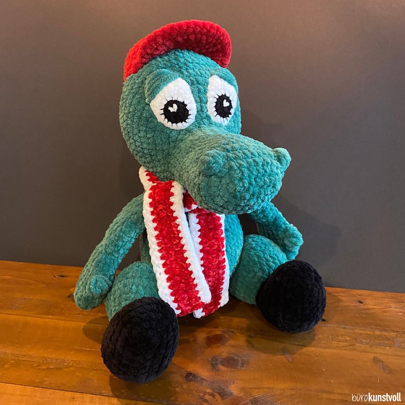 Türkisfarbenes gehäkeltes Krokodil-Amigurumi mit applizierten Augen, roter Mütze und rot-weißem Schal