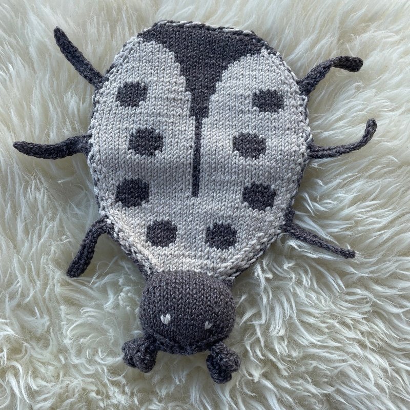 Gestrickter Marienkäfer in Grau und Weiß mit dunklen Punkten und kleinen Herzstichen auf flauschigem Untergrund