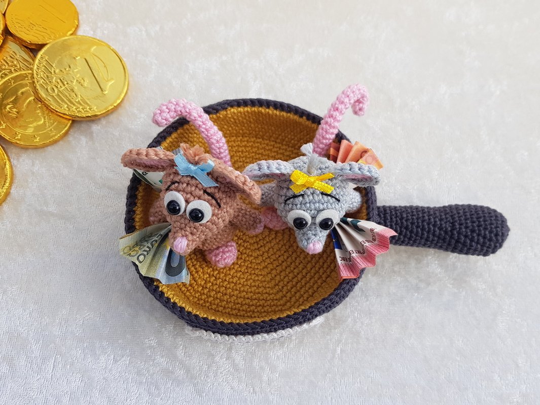 Zwei braune und graue Amigurumi‑Mäuse mit Wackelaugen und Schleifen in einer gehäkelten Pfanne, sie halten gefaltete Geldscheine und sind von goldenen Schokotalern umgeben