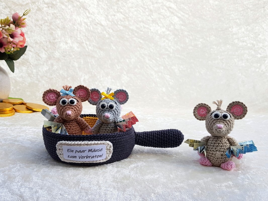 Drei gehäkelte Amigurumi-Mäuse, zwei in einer gehäkelten Pfanne und eine daneben, dekoriert mit gefalteten Papiergeldscheinen und dem Schild 'Ein paar Mäuse zum Verbraten!'