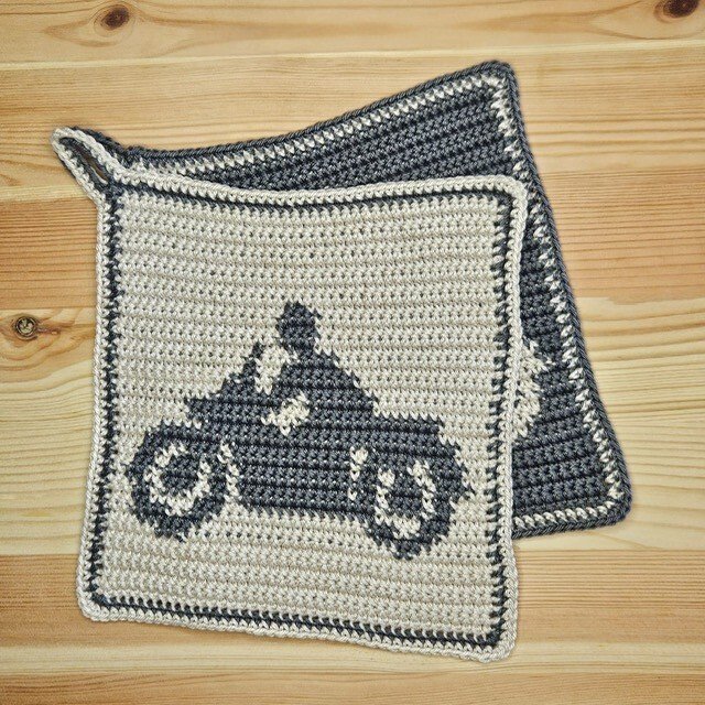 Zwei gehäkelte quadratische Topflappen in beige und grau, einer mit dunkelgrauer Motorrad-Silhouette und kontrastierendem Rand, auf Holzuntergrund