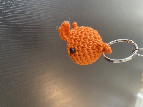 Gehäkelter orangefarbener Fisch-Schlüsselanhänger mit schwarzem Perlenauge und Metallring vor dunklem Hintergrund.