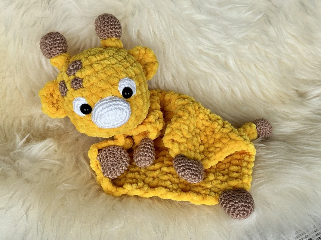 Yellow crocheted giraffe snuggle blanket with brown accents and white snout on a fluffy white surface.