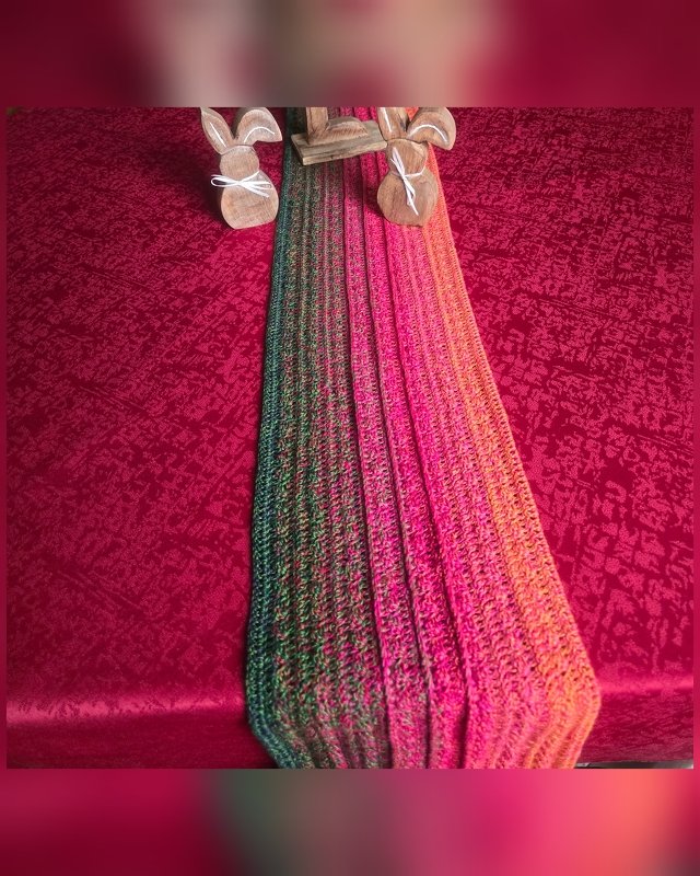 Gehäkelter mehrfarbiger Tischläufer (Grün–Orange–Pink) auf roter Tischdecke mit zwei Holzhasen