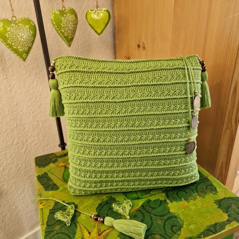 Quadratisches grünes gehäkeltes Kissen mit Quasten, Holzperlen und runden Anhängern auf grün gemustertem Tisch