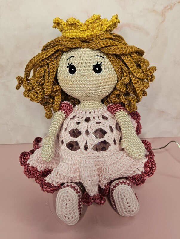 Gehäkelte Amigurumi-Puppe sitzend mit goldener Krone, lockigem Haar und rosafarbenem Rüschenkleid