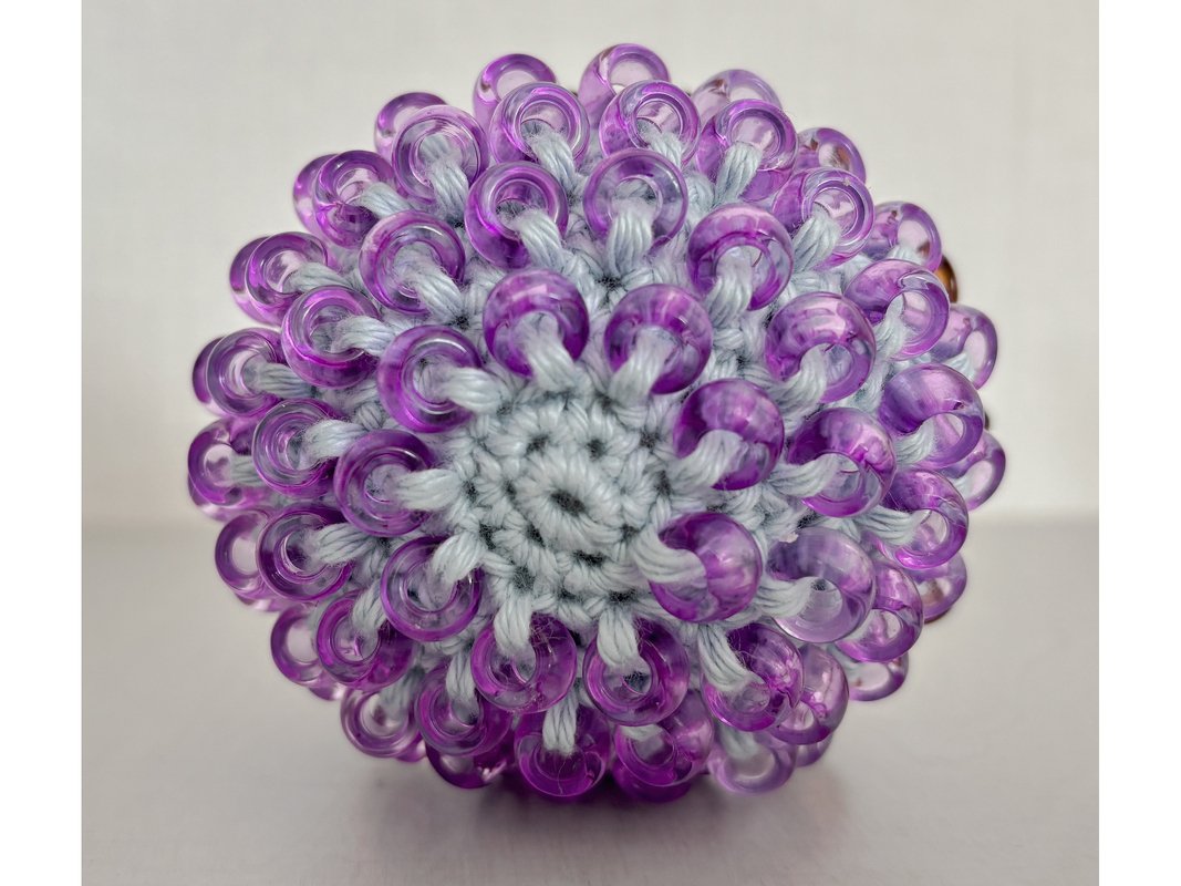 Close-up of a handmade crocheted ball in light grey yarn densely covered with translucent purple beads.
