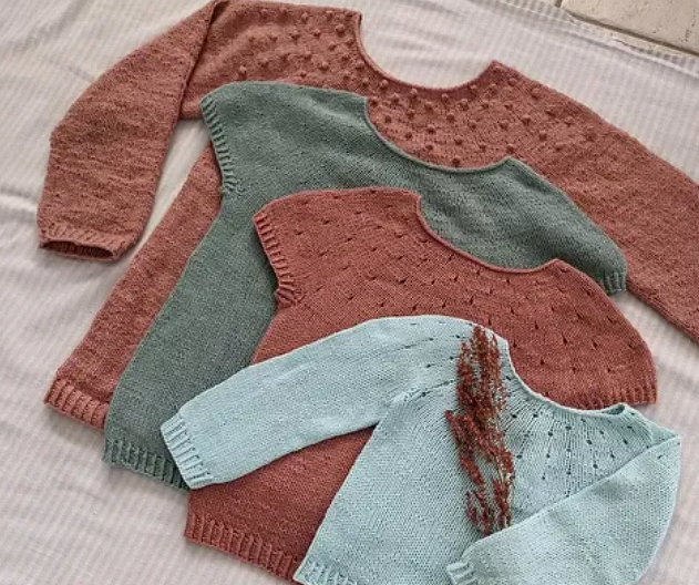 Vier gestrickte Baby- und Kinderpullover in Pastellfarben mit rundem Ausschnitt und punktartiger Struktur, nebeneinander auf hellem Stoff arrangiert.