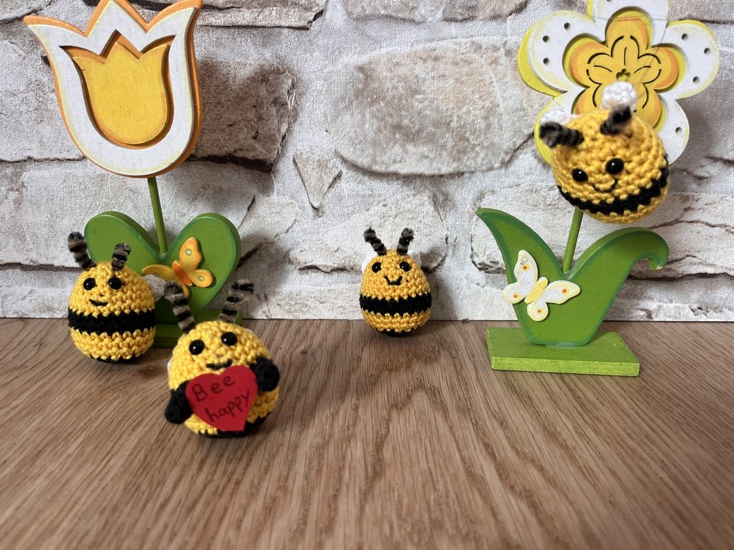 Mehrere gehäkelte Bienen in Gelb und Schwarz, arrangiert mit Holzblumen- und Schmetterlingsdekoration auf einem Holztisch.