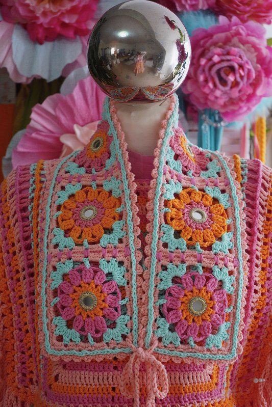 Bunte gehäkelte Weste in Pink, Orange und Türkis mit floralen Granny Squares und kleinen Spiegeleinsätzen auf einer Schaufensterpuppe.