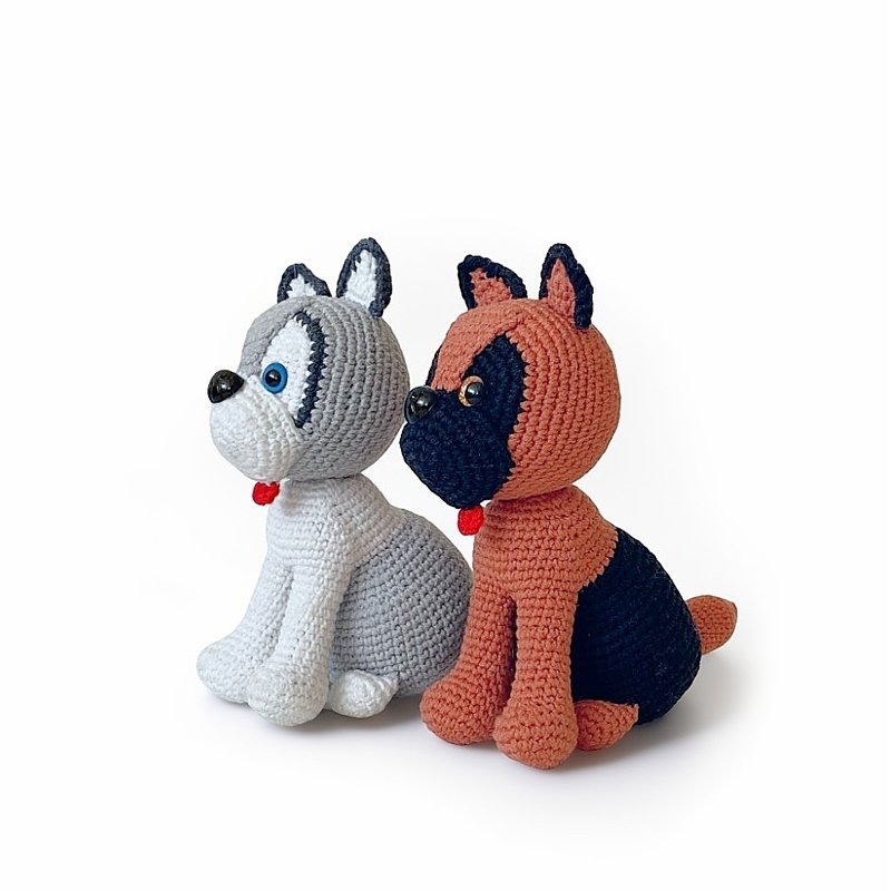Zwei sitzende gehäkelte Amigurumi-Hunde, einer grau mit dunklen Gesichtspartien, einer rostbraun mit schwarzem Gesicht, beide mit kleinem roten Detail am Hals