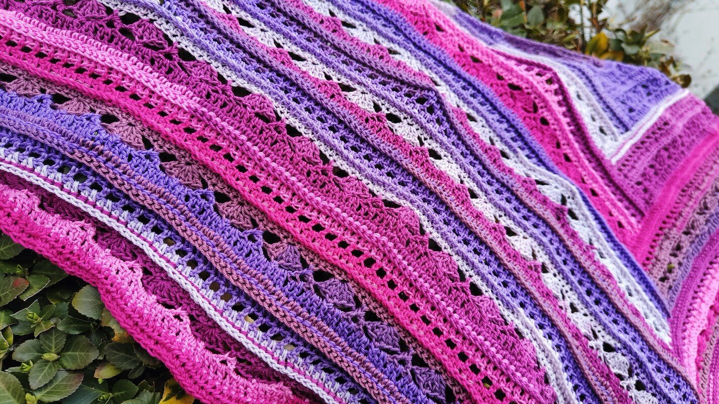 Pink and purple crocheted triangle shawl with lace stripes and scalloped edge draped over foliage