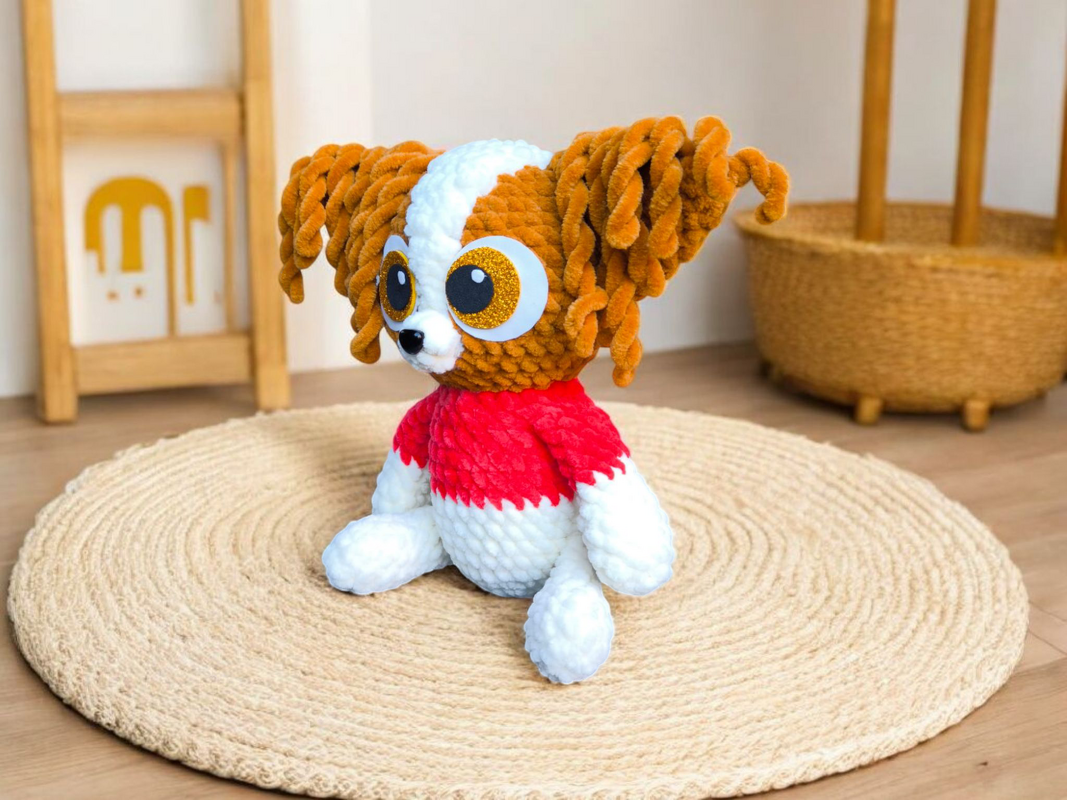 Gehäkelter Amigurumi-Hund in Braun, Weiß und Rot mit großen goldenen Augen auf rundem Teppich.