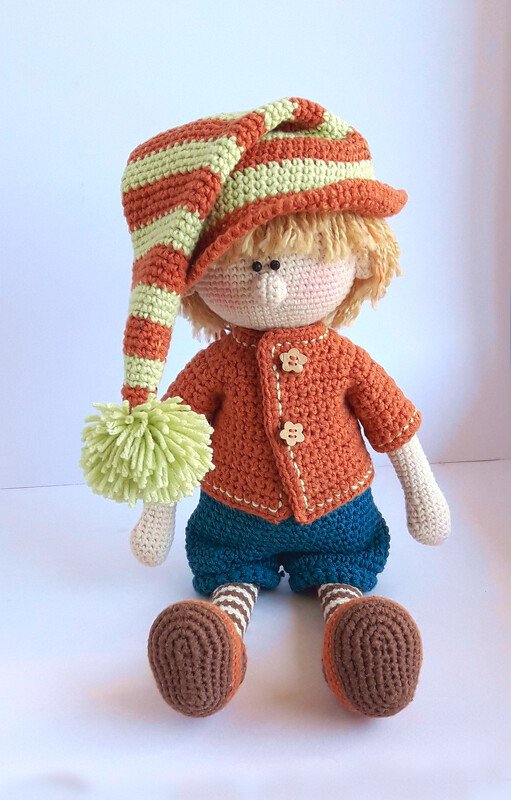 Gehäkelte Amigurumi-Jungenpuppe mit gestreifter Zipfelmütze und Bommel, oranger Jacke, blauer Hose, gestreiften Socken und braunen Schuhen vor hellem Hintergrund.