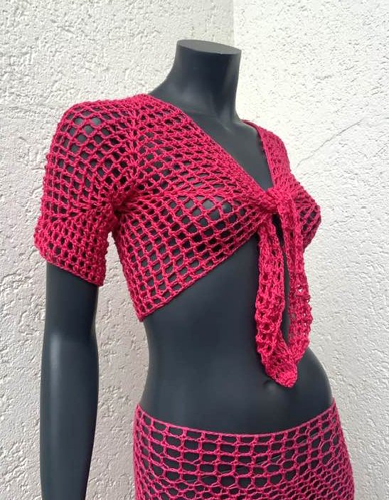Fuchsiafarbener gehäkelter Bolero mit Bindeband und kurzem Netzrock an einer Schaufensterpuppe vor heller Wand.