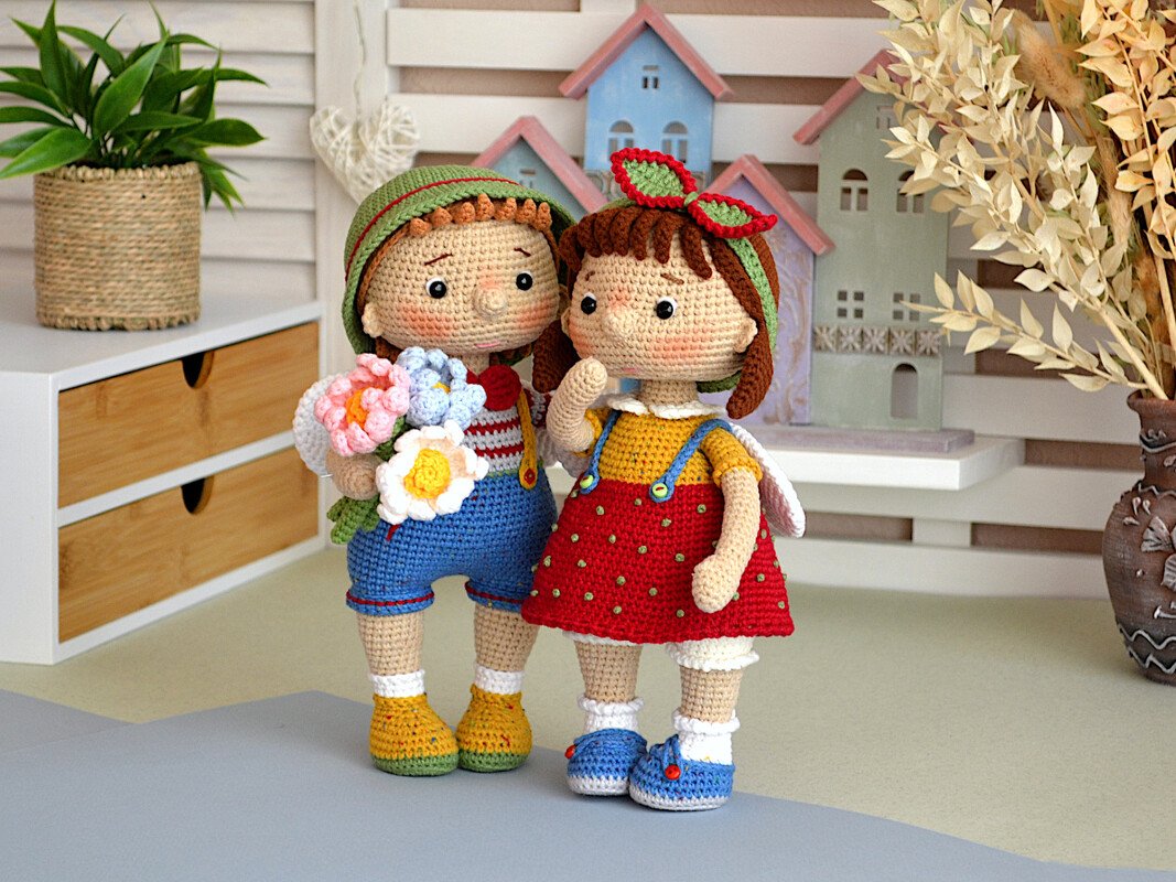 Two crocheted amigurumi dolls, one holding flowers, standing on a tabletop with decorative houses in the background