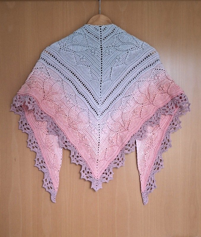 Crocheted triangle shawl with ombre fade from pale grey to soft pink, featuring leaf lace motifs and a scalloped border, hanging on a wooden hanger.